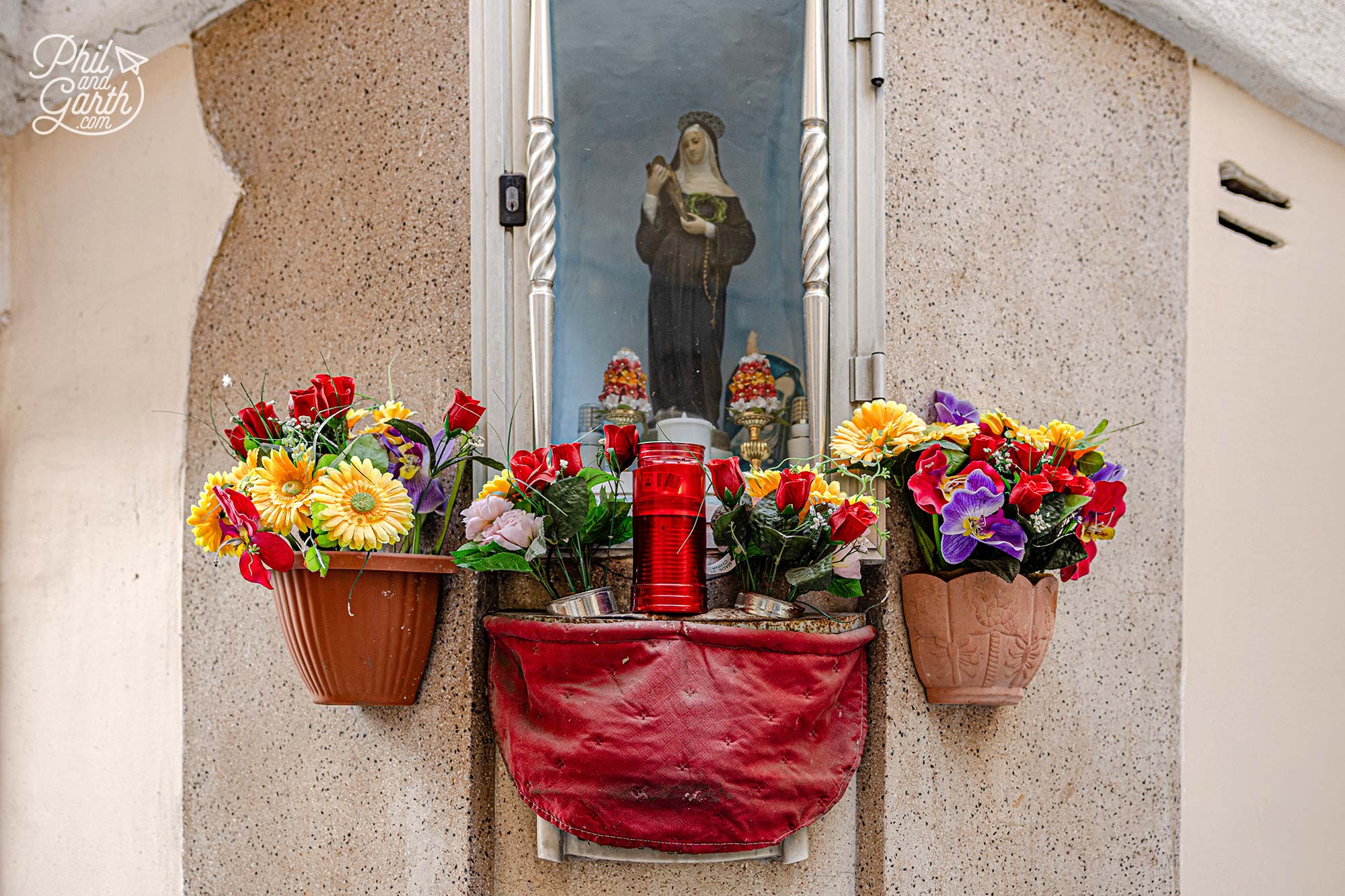 Traditional Maltese street shrine in Valletta featuring a statue of Saint Rita -colourful flowers and devotional candles on a historic limestone building